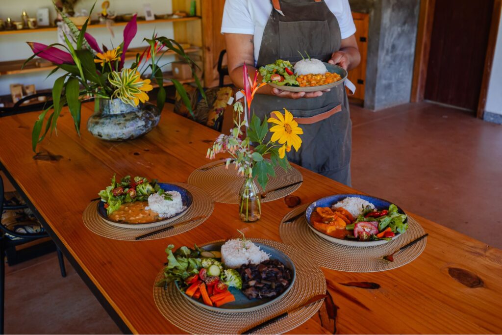 Pratos de refeição saudável e fresca com legumes, arroz e feijão, servidos em mesa de madeira com arranjos florais, destacando a gastronomia da Ecovila Coração da Mata.