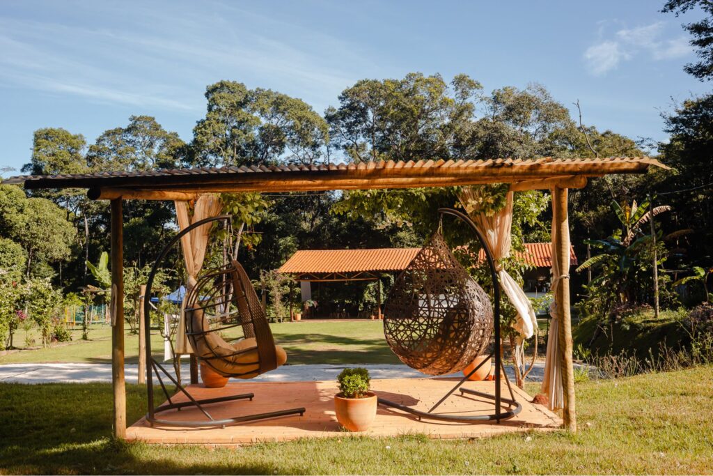 Área de descanso na Ecovila Coração da Mata com pérgola de madeira rústica, redes e cadeiras suspensas em meio à natureza exuberante.