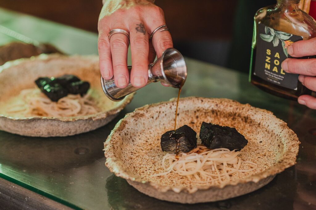 Apresentação criativa de um prato de A Alquimista, com molho escuro e ingredientes frescos, refletindo a fusão de sabores e a culinária com memória afetiva.