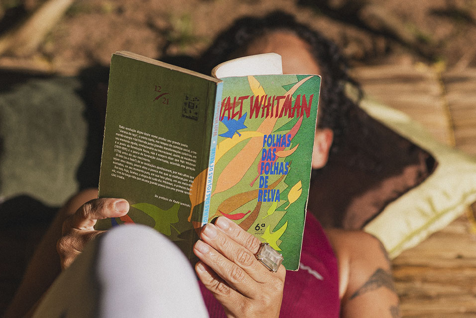 Uma mulher lê livro deitada, apoiada em amolfadas. 