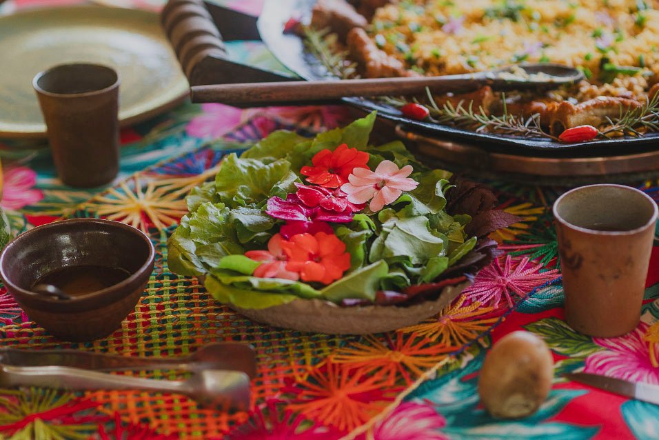 Mesa composta por prato com flores e folhas, cumbuca de copo de barros com refeiçao servida em caçarola. 